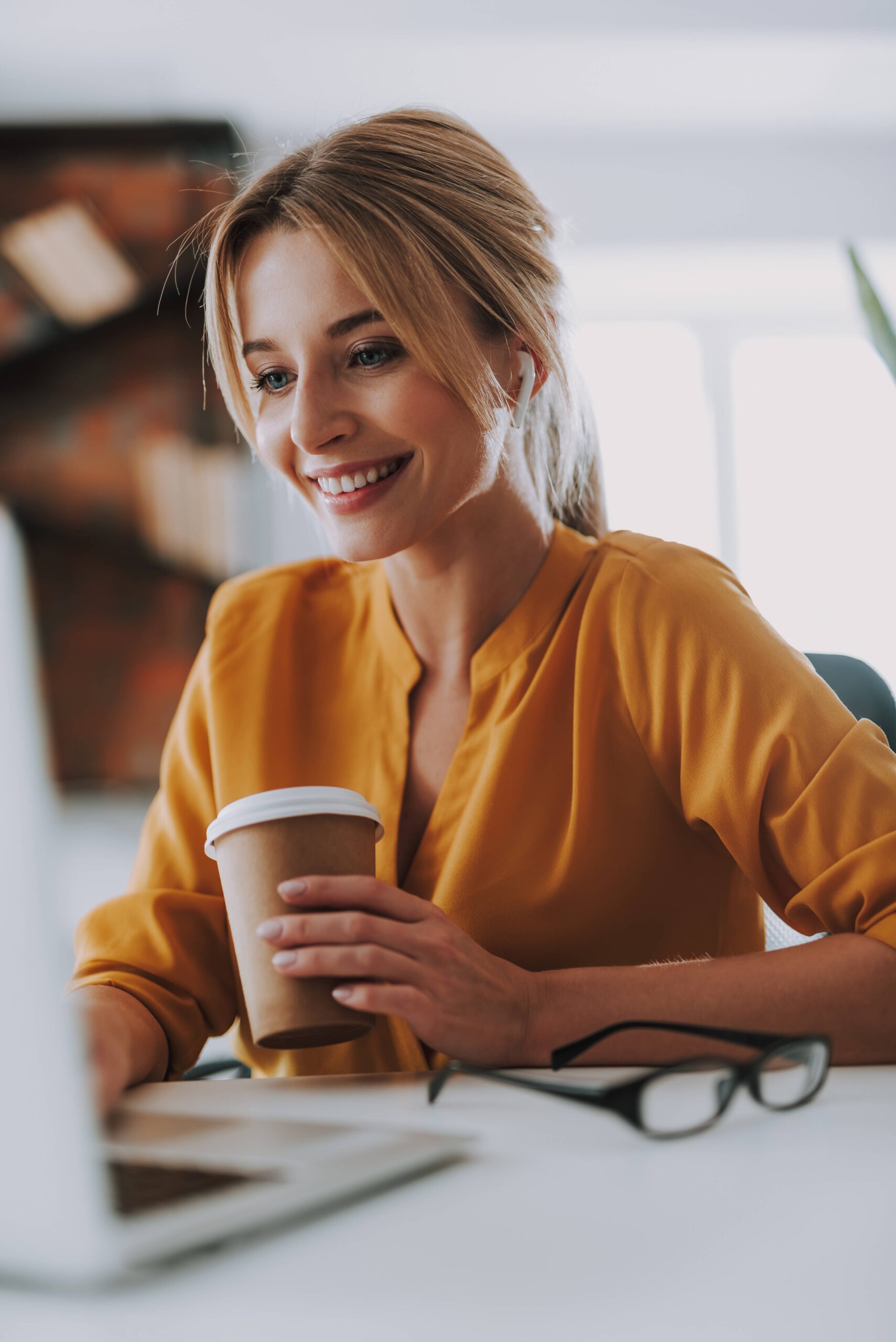 Cheerful young lady working in the office and using wireless earphones while touching the keyboard of her laptop